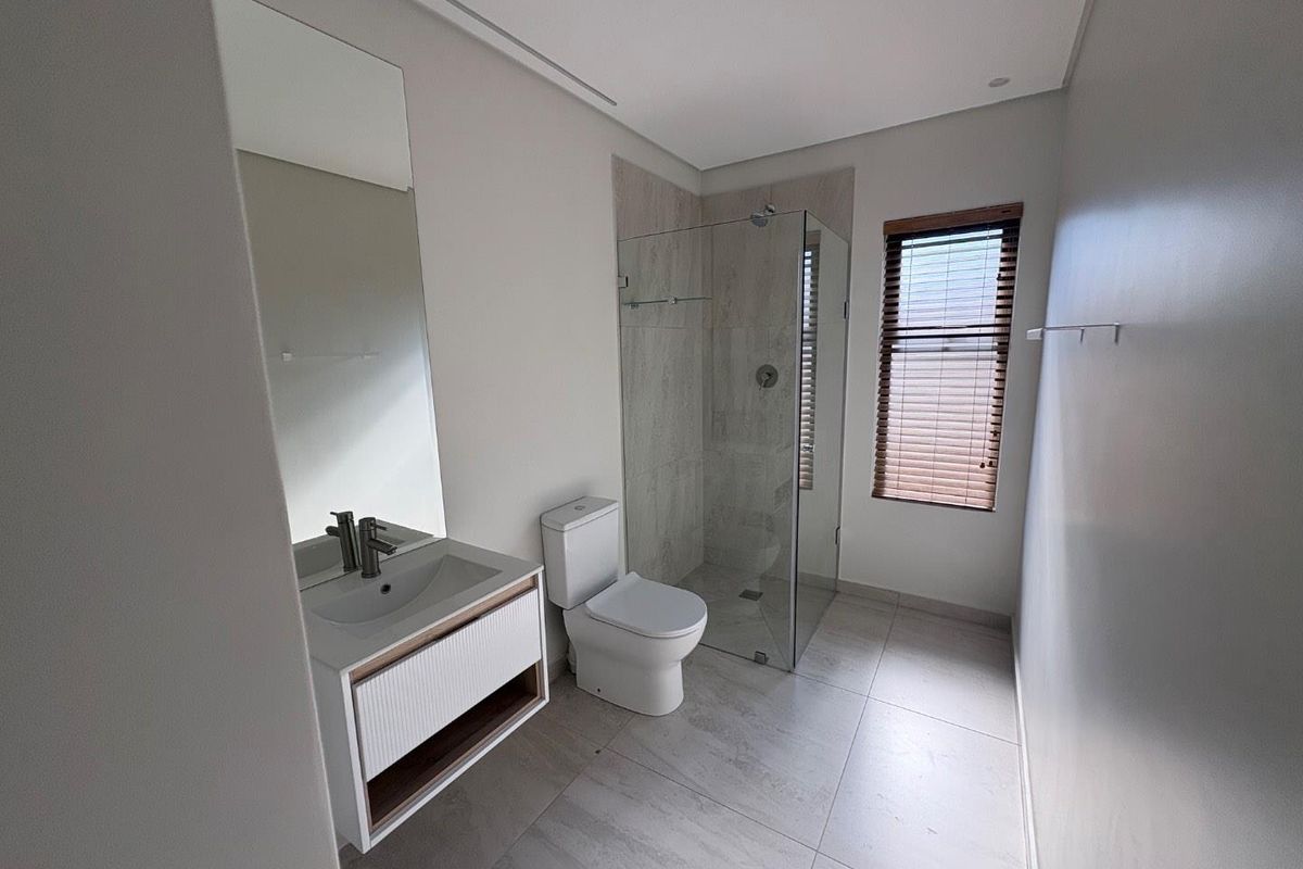 Modern bathroom featuring a sleek vanity, glass-enclosed shower, neutral floor tiles, and a window with fitted blinds, presenting a clean and contemporary aesthetic.