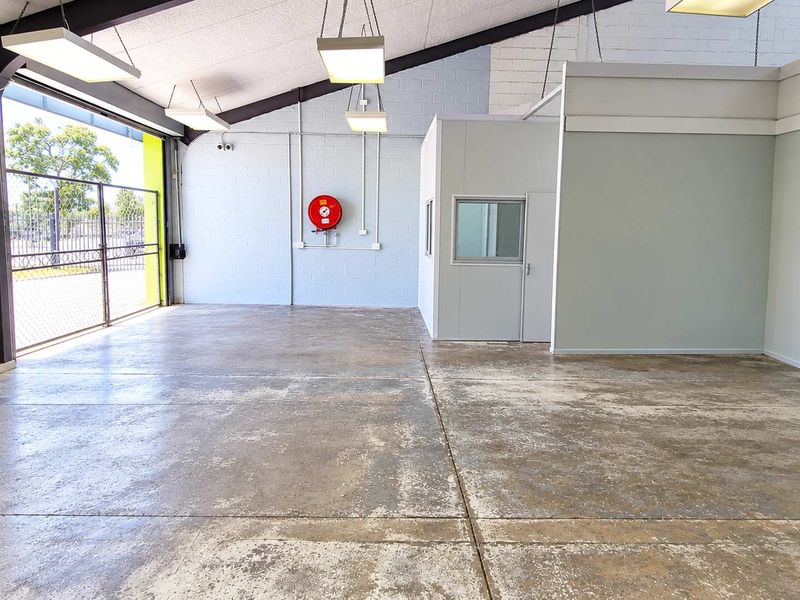 Spacious industrial utility area featuring concrete flooring, exposed steel beams, suspended lighting, and a partitioned office enclosure with a roller shutter door.