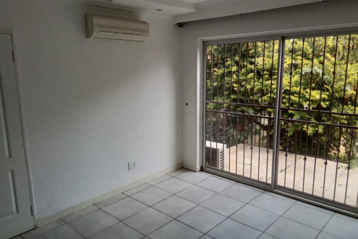 A bright, empty bedroom featuring tiled flooring, a wall-mounted air conditioning unit, and sliding glass doors opening onto a balcony with security bars.