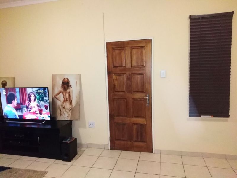 A living room area featuring tiled flooring, a solid wooden door, and dark venetian blinds, providing a functional and neutral living space.