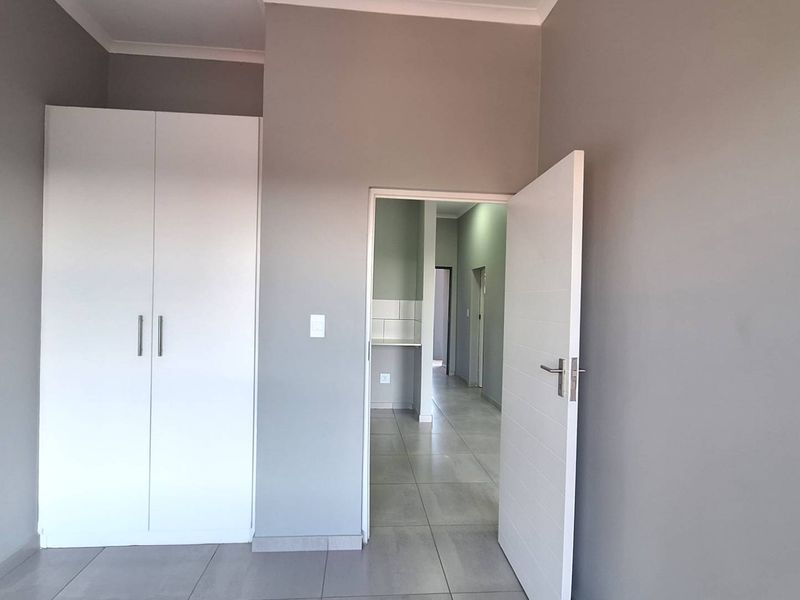 A clean, neutral-toned bedroom featuring built-in wardrobes, tiled flooring, and a view through to the adjacent hallway and kitchen area.