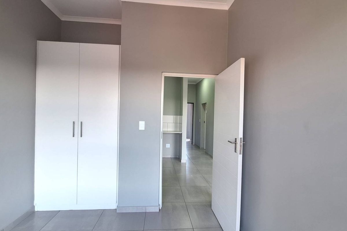 A clean, neutral-toned bedroom featuring built-in wardrobes, tiled flooring, and a view through to the adjacent hallway and kitchen area.