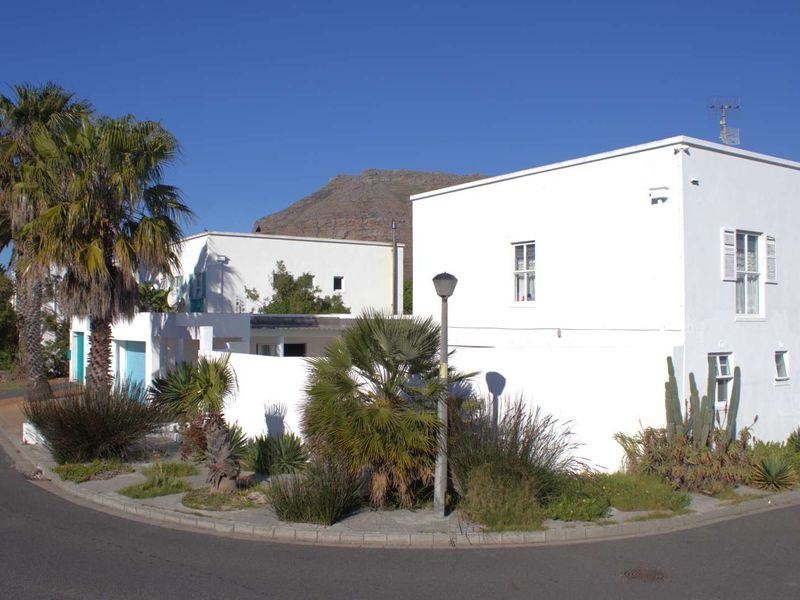 A street-level view of a white-rendered residential property featuring a corner lot position, established landscaping, and clear mountain views in the background.
