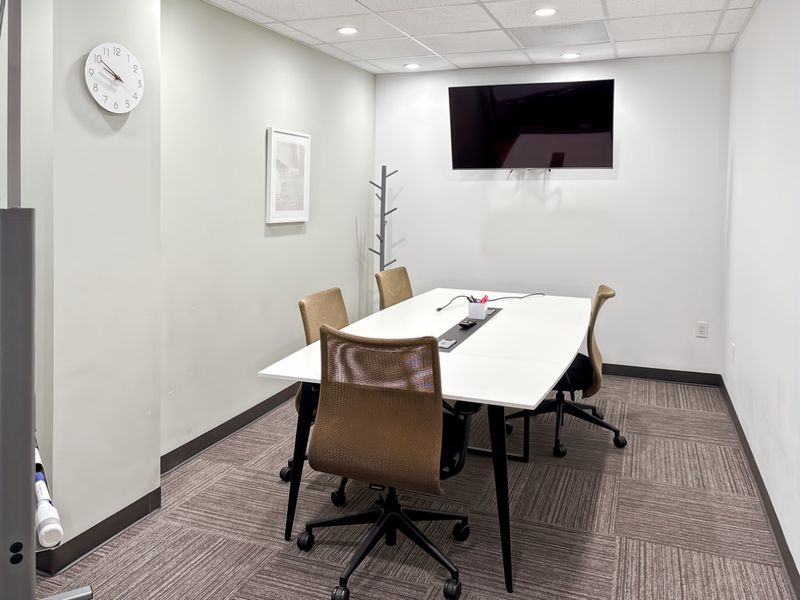 A professional office meeting room featuring carpet tiles, recessed ceiling lighting, and a wall-mounted television for presentations.