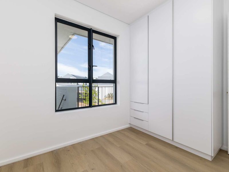 A bright, modern bedroom featuring neutral laminate flooring, clean white walls, and ample built-in cupboard storage with a view through the window.