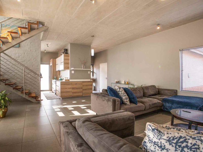 Spacious open-plan living area featuring a modern staircase with stainless steel balustrade, exposed concrete ceilings, and integrated kitchen cabinetry with tiled flooring throughout.