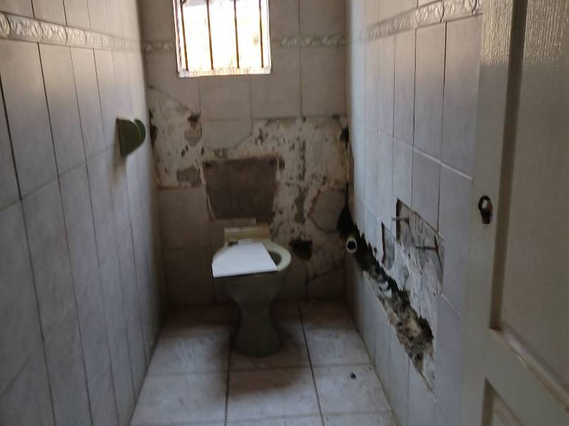 A small bathroom in need of significant renovation, featuring damaged wall and floor tiles and exposed plumbing.