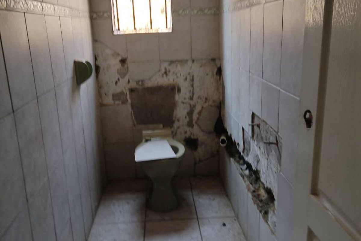 A small bathroom in need of significant renovation, featuring damaged wall and floor tiles and exposed plumbing.