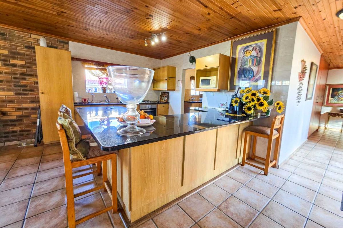 Spacious kitchen featuring a large granite breakfast bar, built-in oven, hob, timber-clad ceiling, and tiled flooring with a rustic brick feature wall.