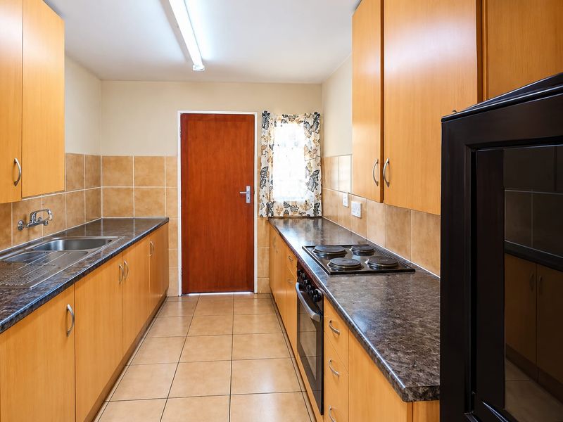 A functional galley-style kitchen featuring light wood cabinetry, dark laminate countertops, built-in oven and hob, and tiled finishes throughout.