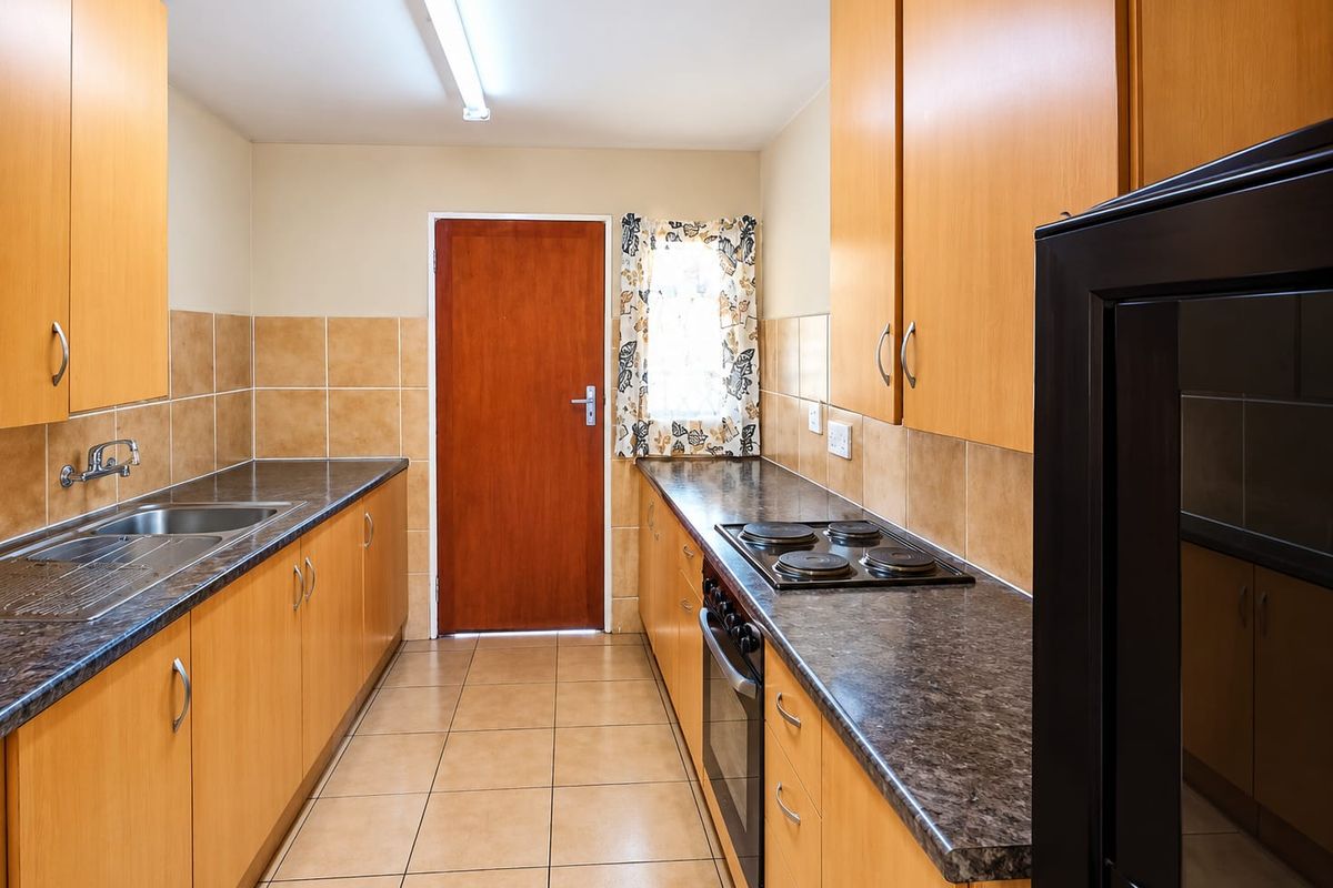 A functional galley-style kitchen featuring light wood cabinetry, dark laminate countertops, built-in oven and hob, and tiled finishes throughout.