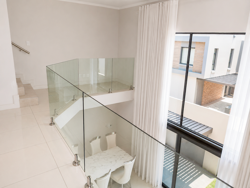 A bright, modern double-volume landing area featuring a glass balustrade, tiled flooring, and large windows overlooking the exterior.