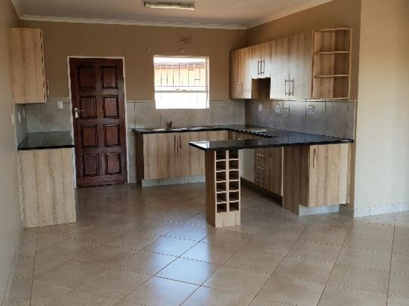 A spacious, empty kitchen featuring light wood-finish cabinetry, dark countertops, a built-in wine rack, and neutral tiled flooring throughout.