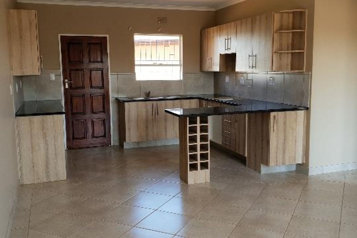 A spacious, empty kitchen featuring light wood-finish cabinetry, dark countertops, a built-in wine rack, and neutral tiled flooring throughout.