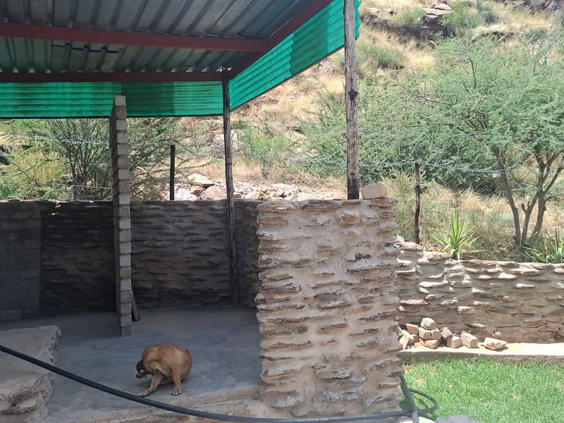 An outdoor covered patio area featuring rustic stone masonry walls, a corrugated metal roof, and concrete flooring, set against a natural hillside backdrop.