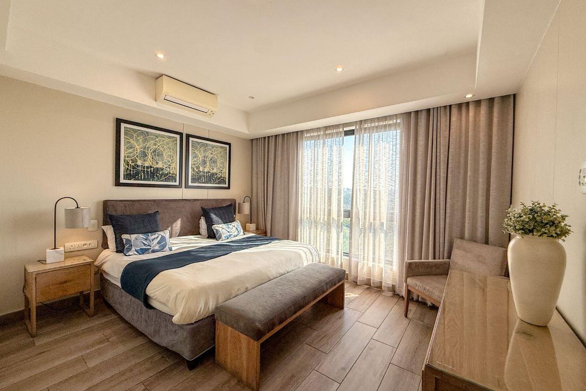 A well-maintained bedroom featuring wood-look tile flooring, recessed ceiling lighting, and a wall-mounted air conditioning unit with large windows and floor-to-ceiling curtains.