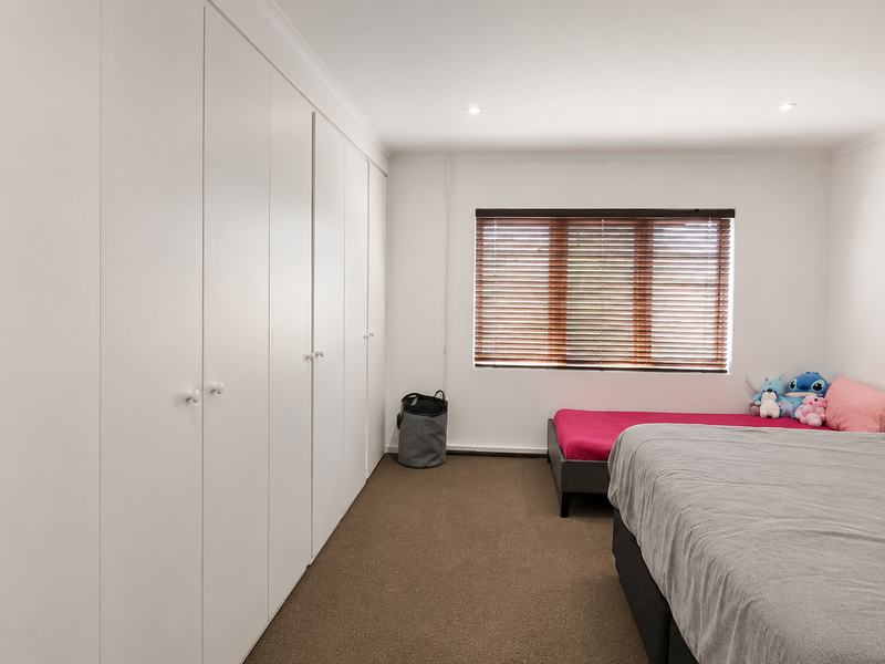 A well-maintained bedroom featuring extensive floor-to-ceiling built-in wardrobes, neutral carpet flooring, and venetian blinds for privacy.