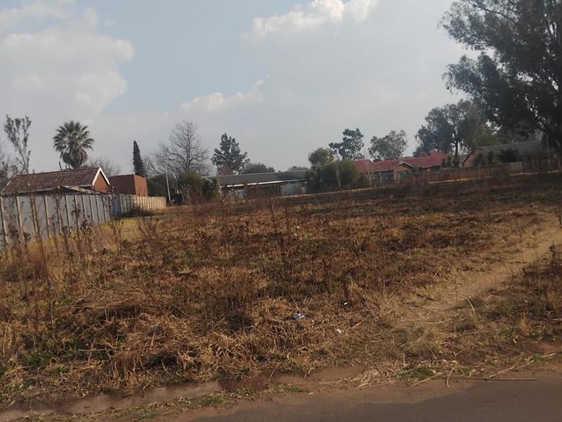A view of a vacant residential plot of land featuring dry vegetation and boundary fencing, viewed from the adjacent street.