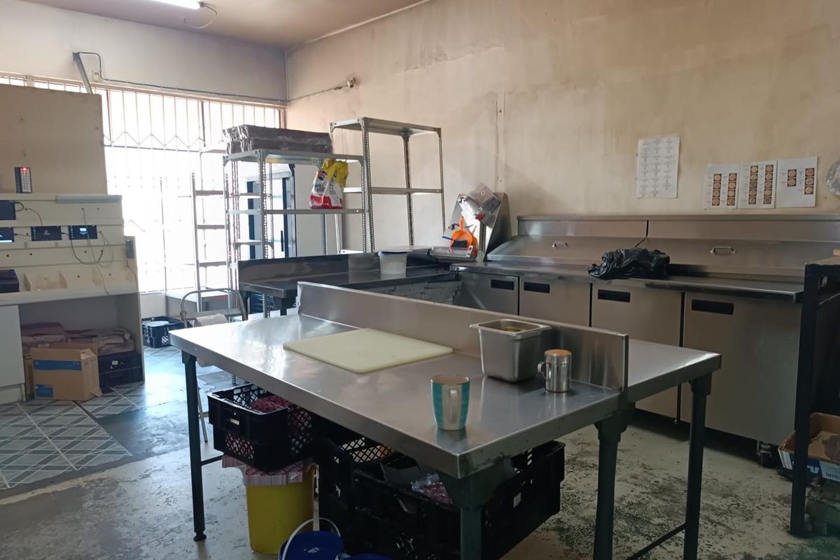 Commercial kitchen space featuring stainless steel prep tables, under-counter refrigeration units, metal shelving, and security-barred windows.