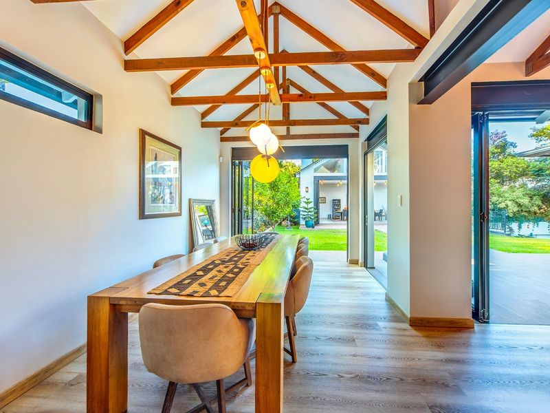 Bright dining area featuring vaulted ceilings with exposed timber trusses, modern laminate flooring, and seamless indoor-outdoor flow through large stacking glass doors.