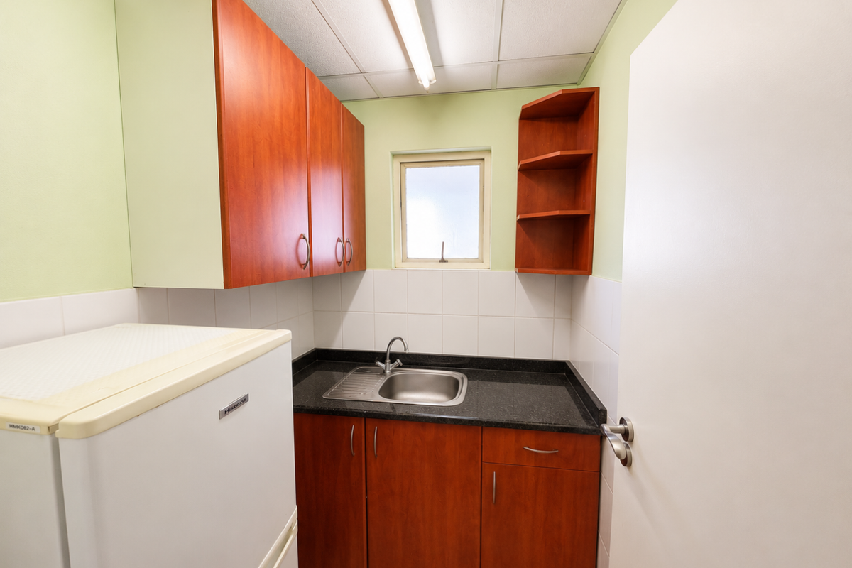 A compact office kitchenette featuring wood-finish cabinetry, a black laminate countertop, a stainless steel sink, and tiled splashbacks.