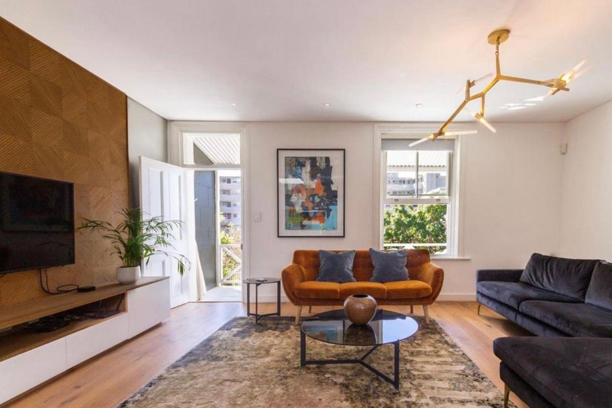 A bright and modern living room featuring timber flooring, a textured feature wall, and large sash windows providing natural light and access to a balcony.