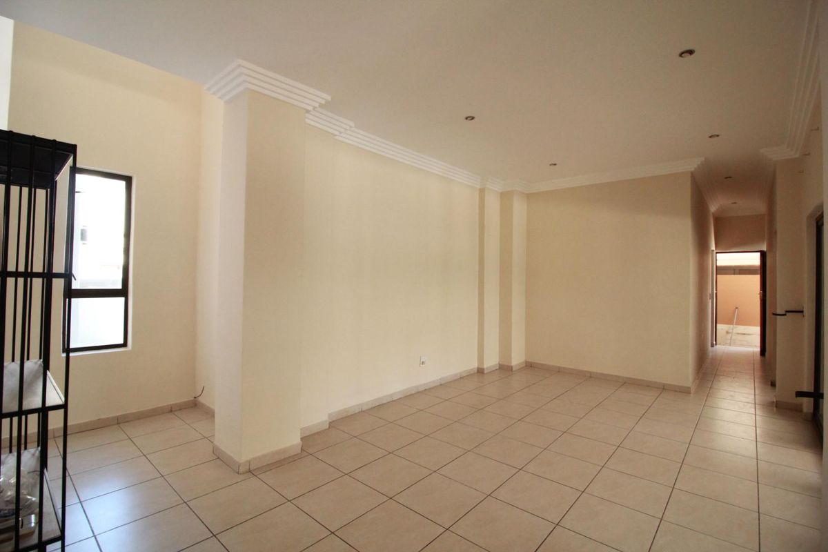 An empty, open-plan living area featuring neutral tiled flooring, recessed downlights, and decorative crown molding, leading to a hallway and exterior access.