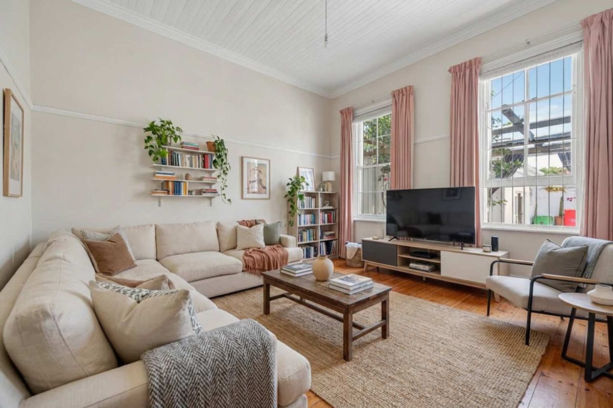 A bright and well-maintained living room featuring original timber flooring, high ceilings with beaded detailing, and large sash windows with security bars.