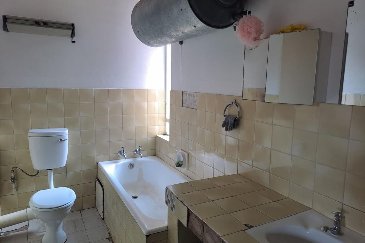 A dated bathroom featuring a built-in bathtub, a tiled vanity with a basin, and a standard toilet, with exposed water heating infrastructure.