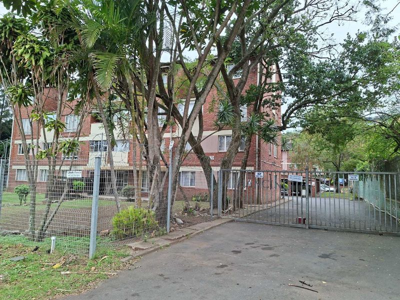 Front view of a multi-story brick apartment building featuring secure gated access, paved driveway, and established surrounding trees.