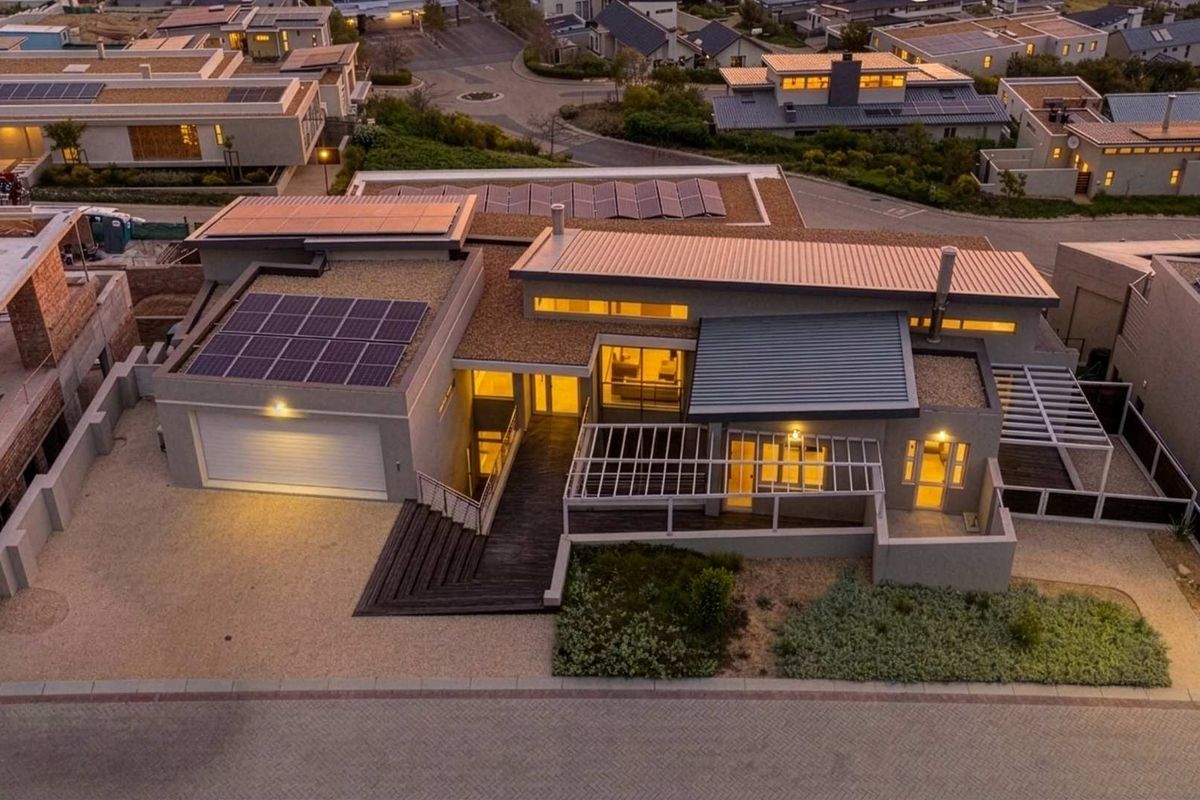 Aerial view of a modern residential property featuring solar panels, a metal roof, timber decking, and a paved driveway in a suburban estate.