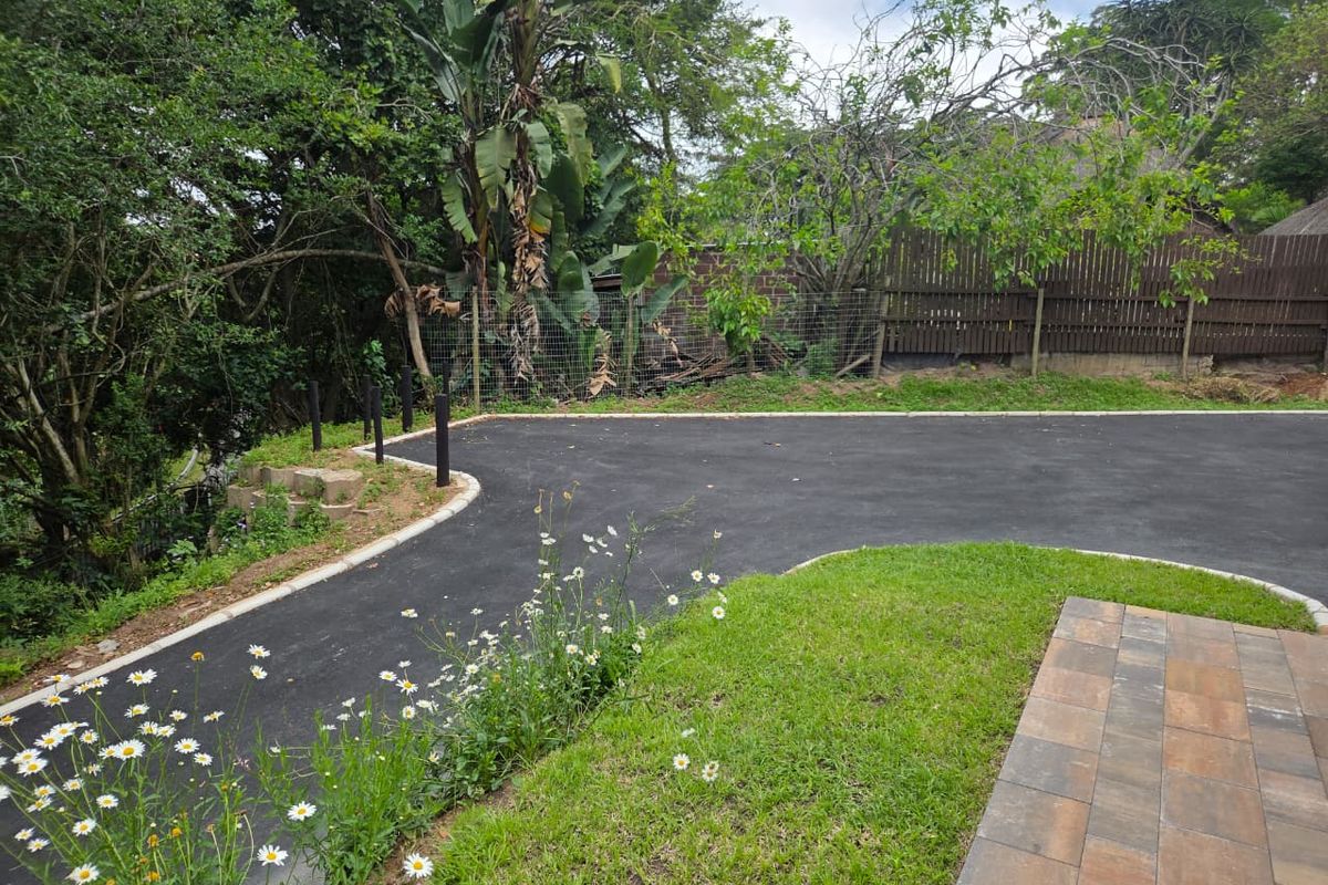 A well-maintained rear outdoor area featuring a paved patio, asphalt driveway, and established garden borders with perimeter fencing.