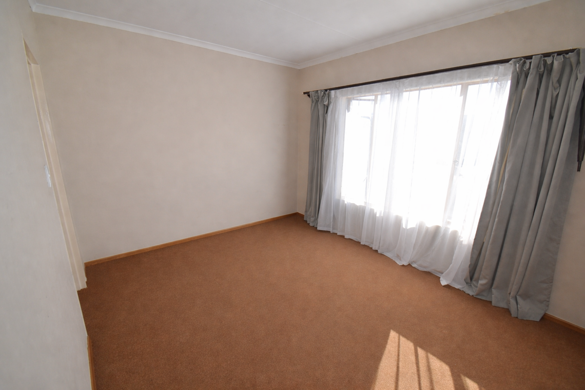 A bright, empty bedroom featuring neutral wall tones, brown carpeting, and large windows with installed curtain tracks.