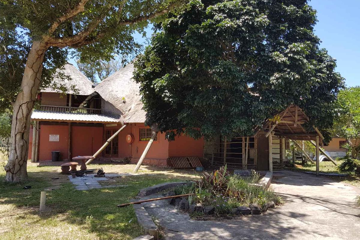 Exterior view of a residential property featuring a thatched roof structure, mature trees, and a paved driveway area.