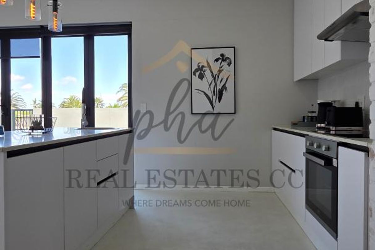 Modern kitchen featuring sleek white cabinetry, integrated oven, extractor fan, and a central island with stone countertops, leading to an outdoor patio area.