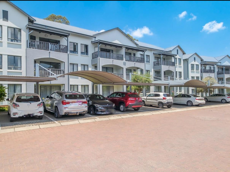 A well-maintained multi-story apartment complex featuring covered parking bays and private balconies for each unit, set within a paved residential area.