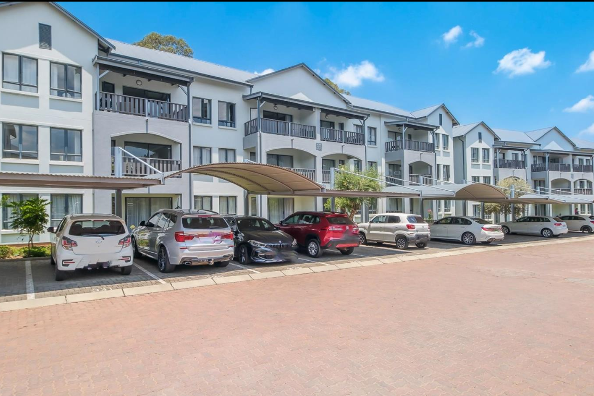 A well-maintained multi-story apartment complex featuring covered parking bays and private balconies for each unit, set within a paved residential area.