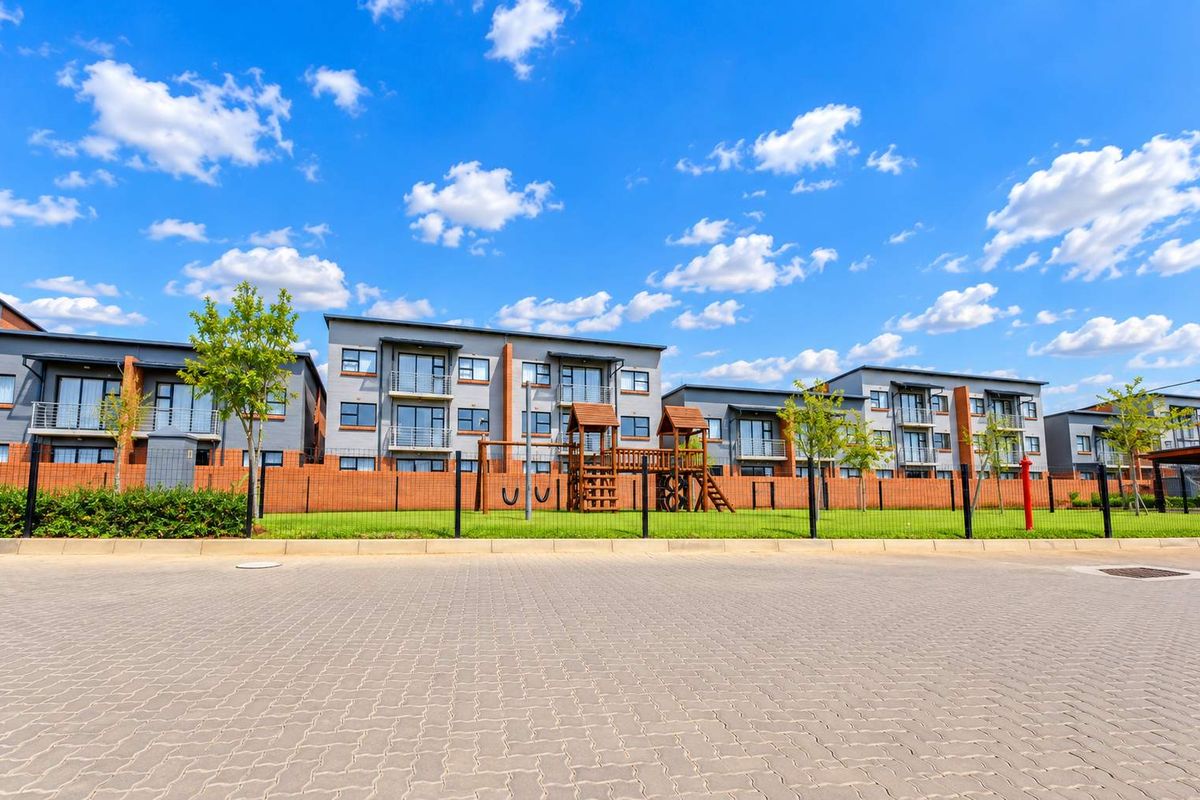 Exterior view of a modern apartment complex featuring paved driveway, brick boundary walls, and a communal playground area.