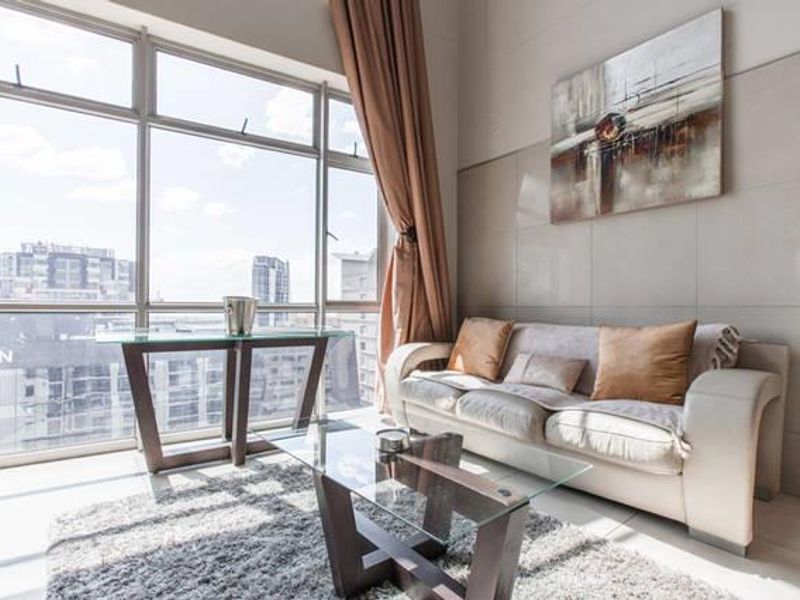 Bright living area featuring large floor-to-ceiling windows with city views, neutral tiled flooring, and a modern tiled feature wall.