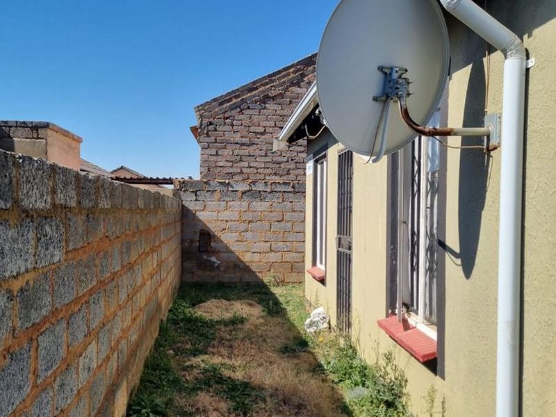 A narrow rear exterior side passage featuring a boundary wall, security bars on windows, and a mounted satellite dish.