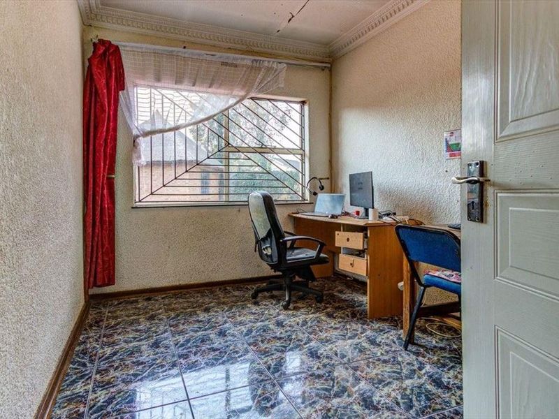 A small office space featuring tiled flooring, security bars on the window, and decorative ceiling cornicing, currently used as a workspace.