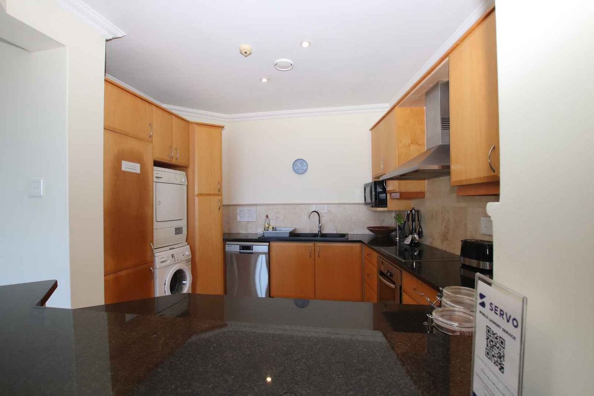 A functional kitchen featuring wooden cabinetry, dark granite countertops, integrated appliances, and a built-in laundry area with stacked washer and dryer.