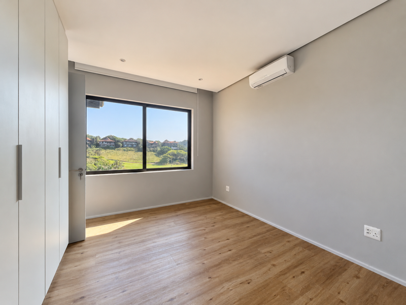 A bright, modern bedroom featuring light wood-look laminate flooring, floor-to-ceiling built-in cupboards, a split-system air conditioner, and scenic views through a large window.