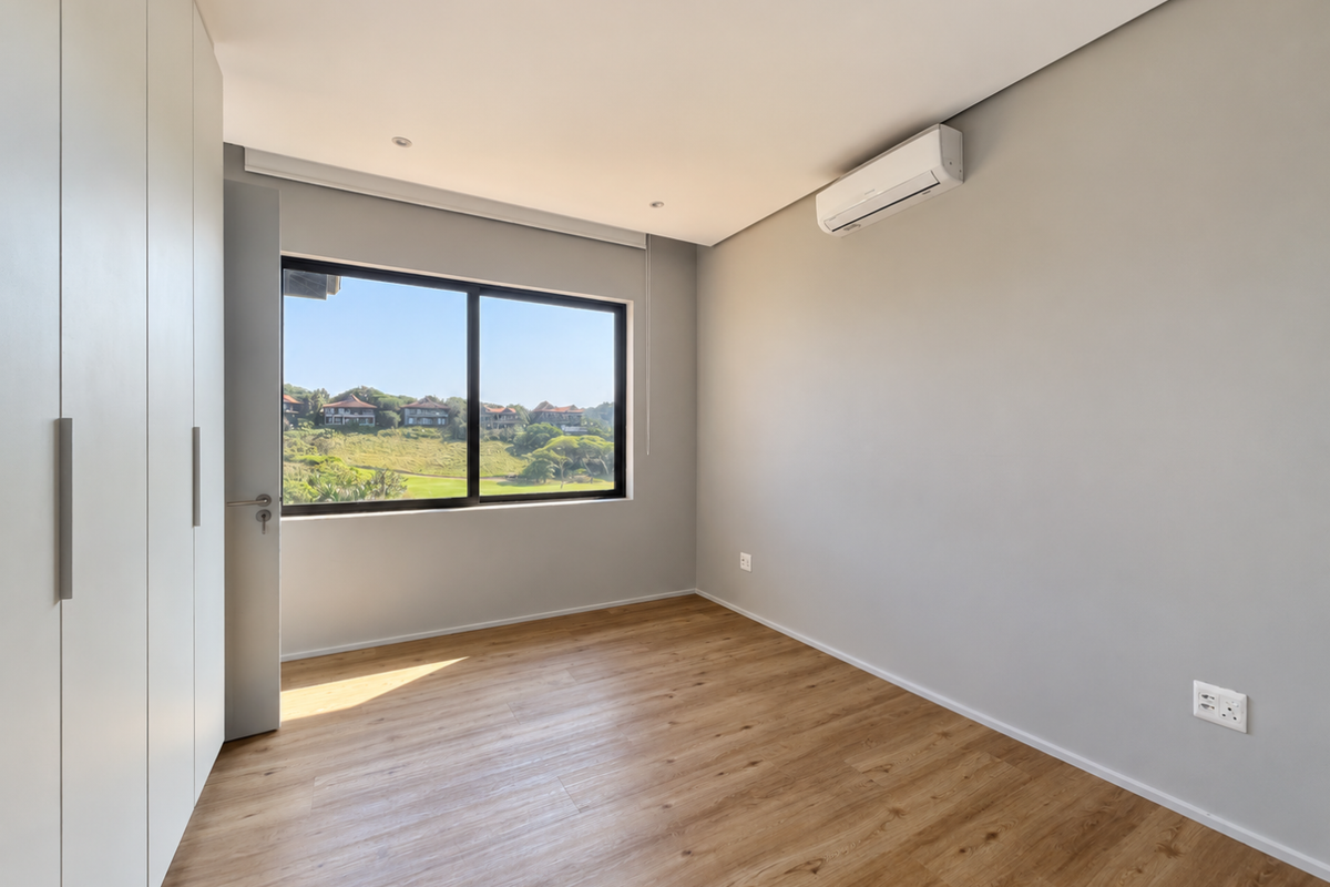 A bright, modern bedroom featuring light wood-look laminate flooring, floor-to-ceiling built-in cupboards, a split-system air conditioner, and scenic views through a large window.