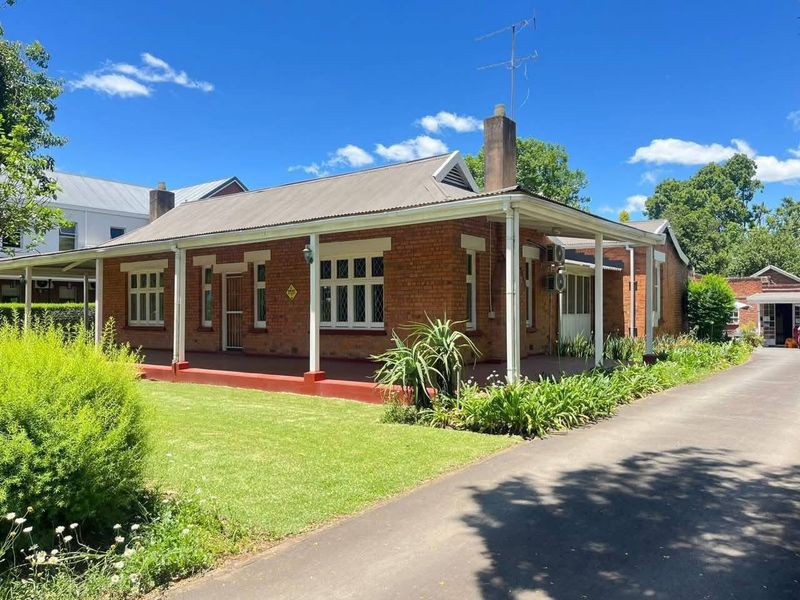 A charming single-story brick office building featuring a covered veranda, corrugated metal roof, and an adjacent asphalt driveway set within a landscaped garden.