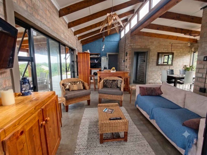 Spacious open-plan living area featuring exposed brick walls, high vaulted ceilings with timber beams, polished concrete floors, and large sliding doors opening to views.