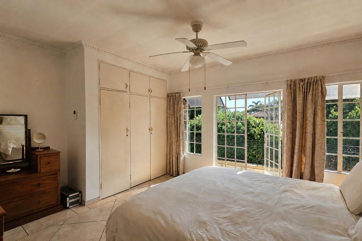 A bright bedroom featuring built-in cupboards, a ceiling fan, and double doors opening onto a private balcony with garden views.