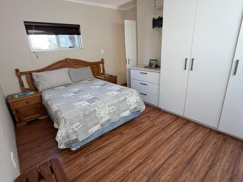 A functional bedroom featuring laminate flooring, built-in white cupboards, and a window with a fitted blind.