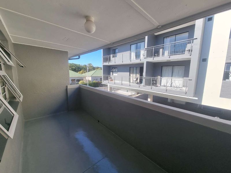 A clean, covered balcony with a concrete floor and metal railing, offering an open view of the adjacent apartment building complex.