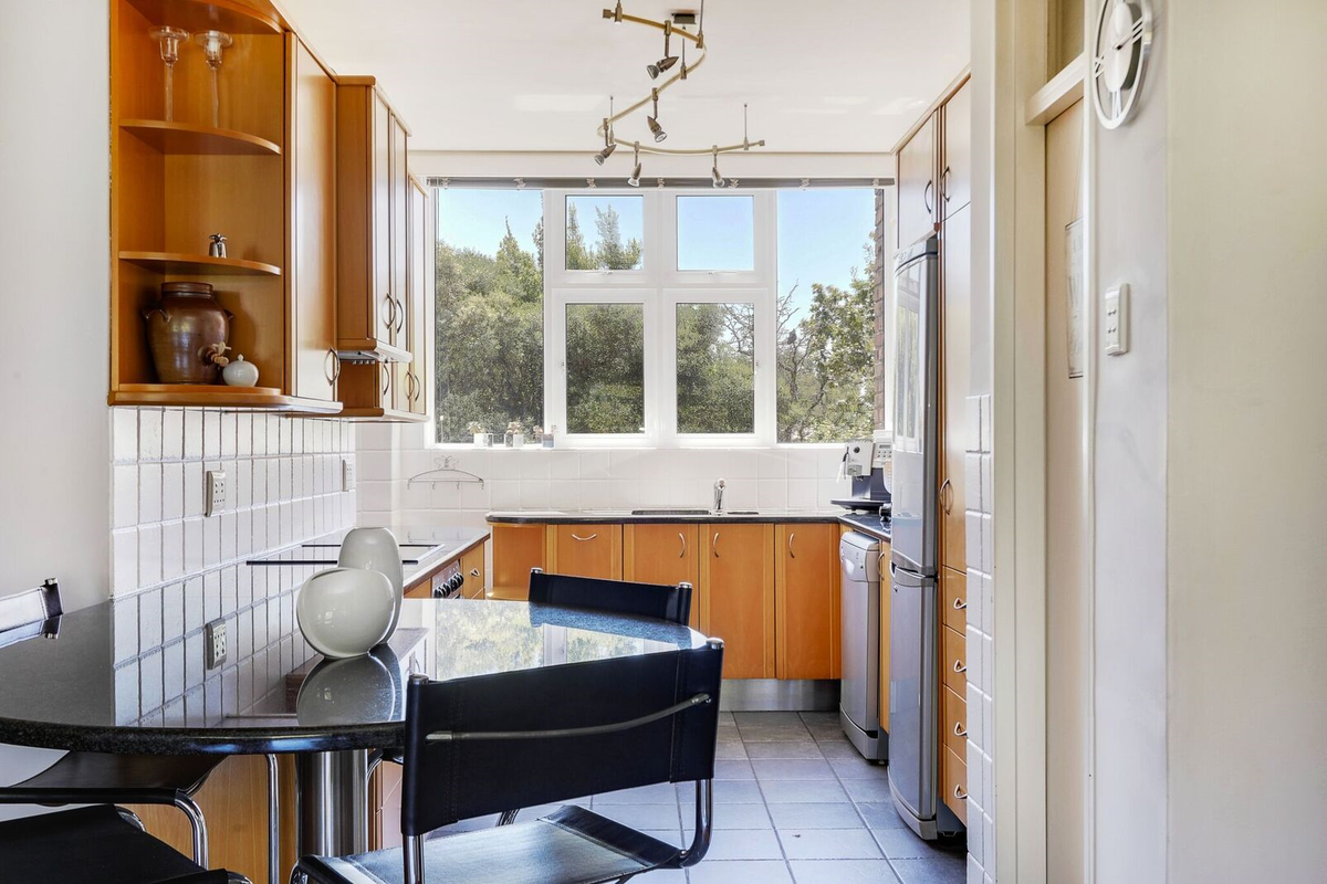 A functional kitchen featuring wooden cabinetry, tiled backsplash, and tiled flooring, with a large window providing natural light and a view of greenery.